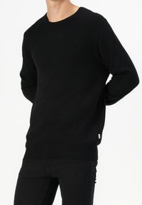 Black knit sweater with a rounded neckline, long sleeves, and a fitted hem. Features a small brand tag on the lower side.