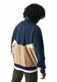 Fleece-Pullover, marinblau mit beige und weißen Einsätzen, hoher Kragen, gerippter Saum, lockere Passform, strukturiertes Material, mit Logo am Hals akzentuiert.