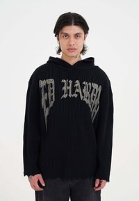 Felpa nera a coste con testo grafico argentato sul davanti, caratterizzata da una vestibilità ampia, cappuccio con cordino e dettagli orlo sfilacciato.