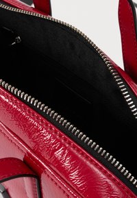 Sarkana spīdīga soma ar sudraba rāvējslēdzēju daļēji atvērta, rādot melnu auduma iekšpusi un sarkanas rokturus.