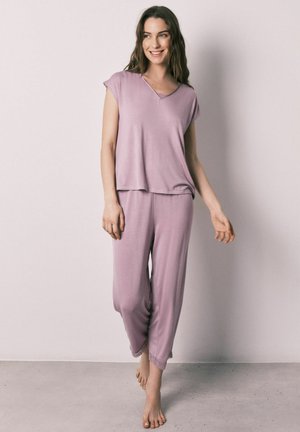 Kvinde med langt brunt hår smilende, gående barfodet, iført en løs lavendel kortærmet top og matchende vidde bukser mod en ensfarvet væg.