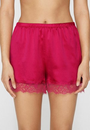 Frau trägt hellpinkfarbene Satinshorts mit elastischem Bund und Spitzenbesatz am Saum, gezeigt von der Taille bis zur Mitte des Oberschenkels.