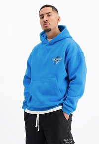 Blauwe hoodie met een voorzak, voorzien van een logo op de linkerkant, gedragen over een witte gelaagde shirt en zwarte shorts.