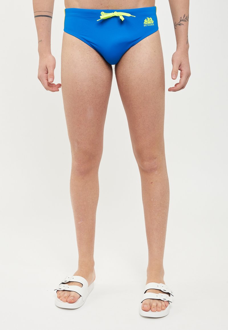 Costumi da bagno blu con cordino giallo e logo, caratterizzati da una vestibilità aderente e una consistenza liscia. Sandali bianchi con doppi cinturini e un leggero plateau.