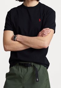 Man met gekruiste armen, gekleed in een zwart T-shirt met rode logo, groene broek met zwarte riem en een horloge met een bruine band.