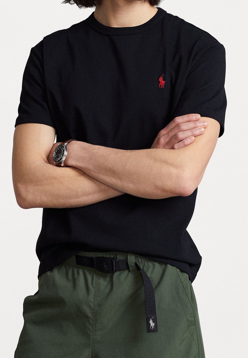 Man met gekruiste armen, gekleed in een zwart T-shirt met rode logo, groene broek met zwarte riem en een horloge met een bruine band.