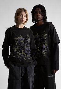 Két ember áll egymás mellett egy sima háttér előtt, fekete réteges koponya grafikás pólókat és sötét nadrágot viselnek.
