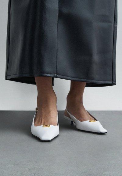Bílé lodičky na podpatku se zavazováním zezadu, špičatými špičkami, zlatými ozdobami, nízkými podpatky a hladkou texturou, kombinované s černou koženou sukní.