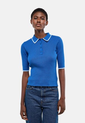 Femme portant un polo bleu côtelé à manches courtes ajusté avec bordures blanches, associé à un jean bleu taille haute, debout devant un fond blanc.