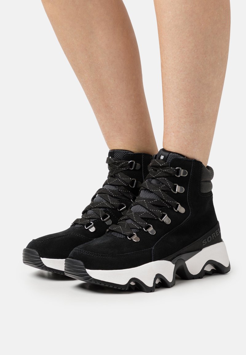 Schwarze Wildleder-Stiefeletten verfügen über ein strukturiertes Obermaterial, metallene Ösen und eine klobige weiße Sohle mit schwarzen Akzenten. Die Schnürdesign verbessert den Sitz.