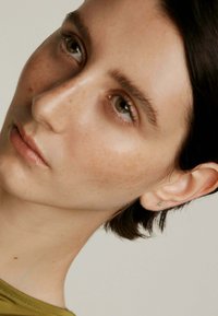 Primer plano de un rostro con piel clara, ligeras pecas, cabello oscuro y lacio, que lleva un pequeño pendiente de aro verde. Look sencillo y natural.