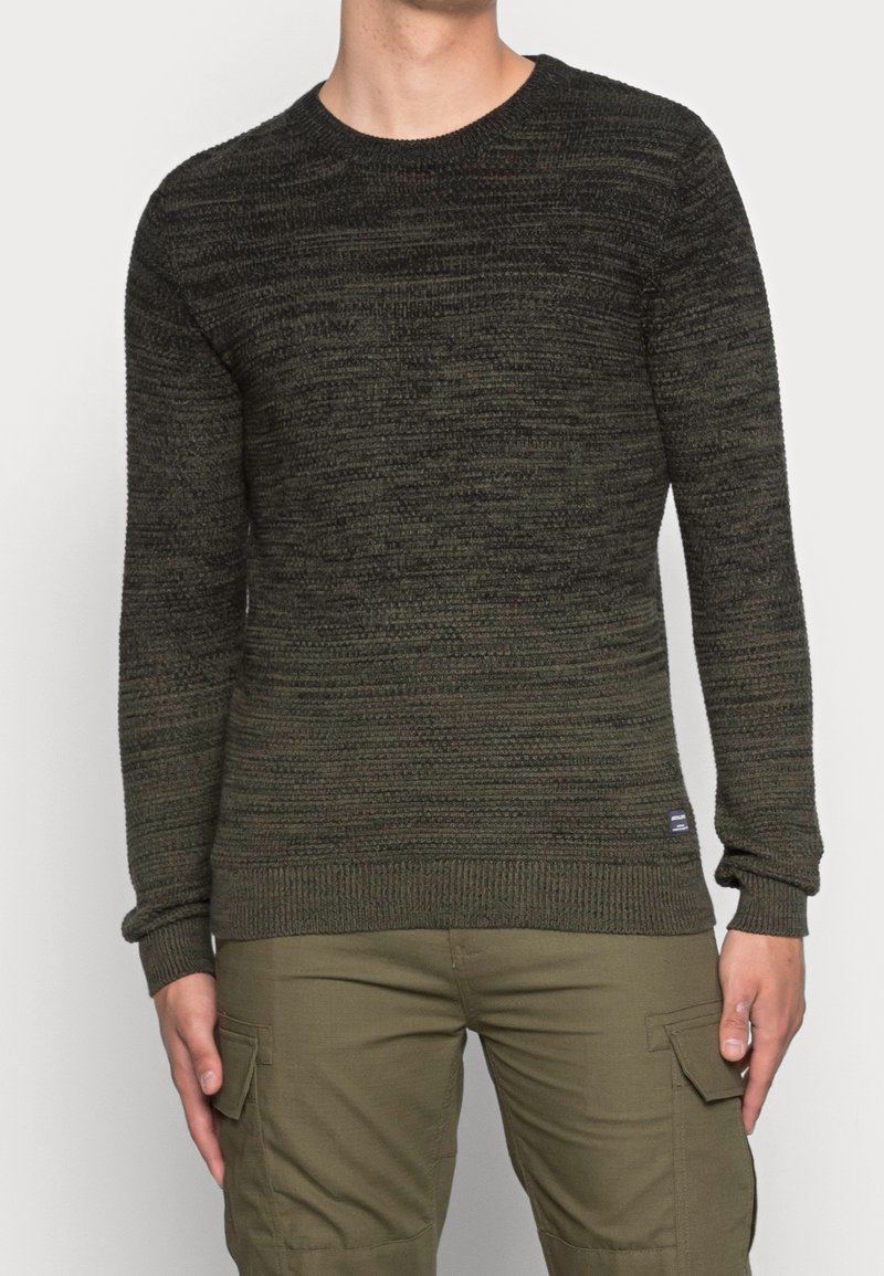 Homme portant un pull en maille chiné vert foncé et noir et un pantalon cargo vert olive, debout devant un fond clair uni.