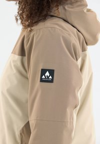 Tan- og beige-jakke i struktureret stof, med et sort firkantet patch på ærmet med et hvidt bjerglogo.