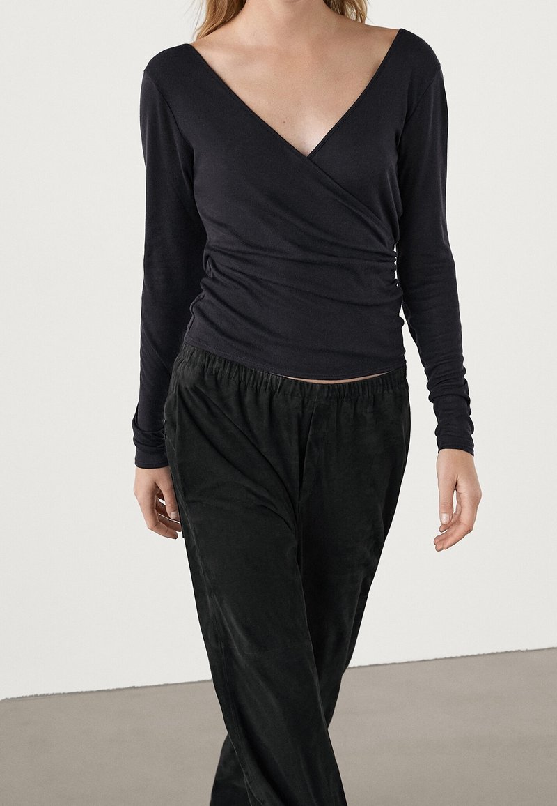 Czarny, długi rękaw, przewiązywany top z dekoltem w kształcie litery V i teksturowanym materiałem, w połączeniu z czarnymi spodniami na elastycznym pasie. Modelka spaceruje po gładkiej powierzchni.
