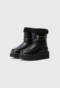 Botas de invierno de cuero negro con suelas de plataforma gruesas, puños de borrego, detalles trenzados y pequeños acentos de cremallera dorada en los lados.