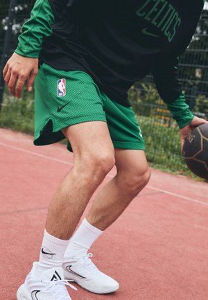 Osoba kozłująca piłkę do koszykówki na zewnętrznym boisku, ubrana w zielone spodenki Celtics, dopasowaną bluzę z długim rękawem, białe skarpetki i białe trampki.