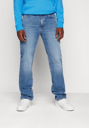 Mann trägt hellblaue Jeans mit geradem Bein, weiße Sneaker und einen leuchtend blauen Hoodie, steht vor einem schlichten hellen Hintergrund.