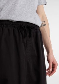 Personne portant un pantalon noir à cordon et un t-shirt gris clair, avec des tatouages textuels visibles sur l'avant-bras.