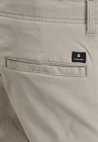 Béžové bavlněné kalhoty s kapsou na zadní straně, s černou čtvercovou etiketou s bílým textem "JACK & JONES". Hladká textura a přizpůsobený design.