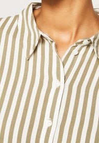 Lyhythihainen, napillinen paita, jossa on pystysuorat raidat oliivinvihreänä ja valkoisena. Kauluksessa on kaulus, ja kankaan pinta on sileä.