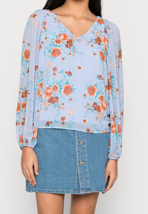 Kvinde iført en lyseblå blomstret bluse med lange ærmer og puffede ærmer samt en blå denim mininederdel med knapper.