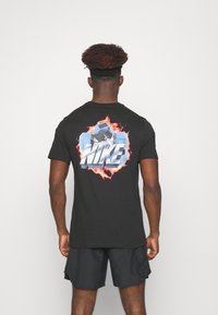 Černé bavlněné tričko s barevnou grafikou váhy a textem "NIKE", umístěné na ohnivém pozadí. Krátké rukávy, design s kulatým výstřihem.