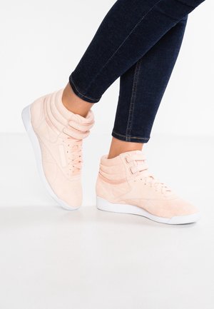 Baskets montantes rose clair avec semelles blanches portées avec un jean skinny bleu foncé, jambes croisées sur un fond blanc.