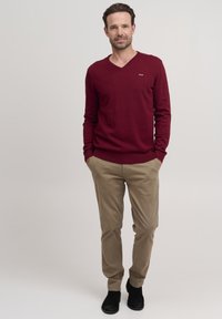 Maglione a maglia bordeaux con scollatura a V, abbinato a pantaloni chinos beige. Il modello indossa scarpe nere, in piedi con le mani nelle tasche.