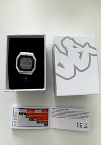 Reloj digital de pulsera con una caja octagonal de plata, acentos en negro y una pantalla digital gris que muestra la hora. Empacado en una caja blanca con el logotipo e instrucciones de cuidado.