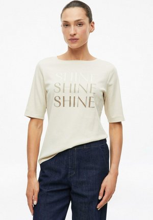 Kobieta z ciemnymi włosami związanymi do tyłu nosi beżowy t-shirt z tekstem ""SHINE"" powtórzonym trzykrotnie oraz ciemnoniebieskie spodnie z wysokim stanem.