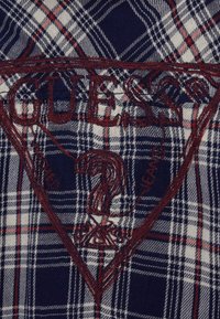 Koszula w granatowo-białą kratę z czerwonym haftowanym logo "GUESS?" w trójkątnym wzorze, z dodatkowymi szczegółami wyhaftowanymi czerwonym nicią.