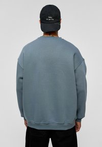 Grijze oversized sweatshirt met een gestructureerde afwerking, geribbelde manchetten en zoom. Zwarte pet met witte tekst op de voorkant. Beide kledingstukken worden achterstevoren gedragen.