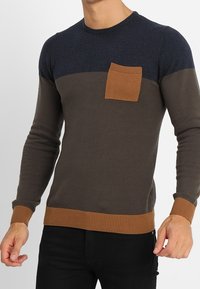 Stickad tröja med en mörkblå överdel, brun torso och ribbad bruna detaljer på kragen och fållen; har en liten bröstficka.