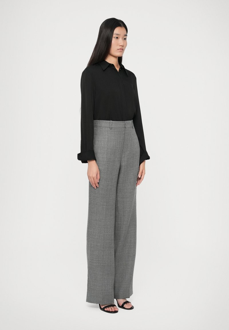 Donna in piedi che indossa una blouse nera a maniche lunghe, pantaloni grigi a gamba larga e sandali neri con punta aperta su uno sfondo bianco semplice.