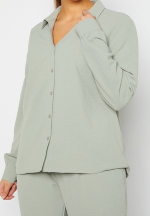 Femme portant une chemise ample vert clair à boutons et un pantalon assorti, tenant le col d’une main.