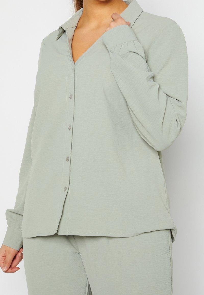 Femme portant une chemise ample vert clair à boutons et un pantalon assorti, tenant le col d’une main.