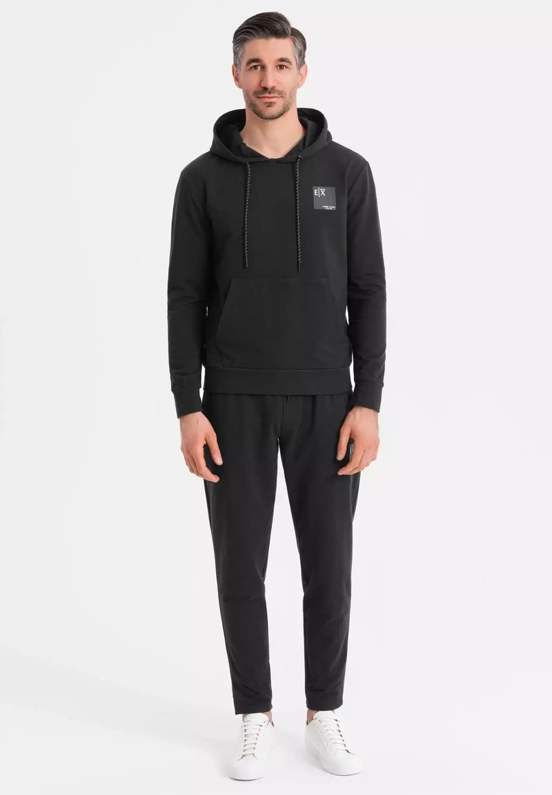 Svart huvtröja med snoddar, känguruficka och logopatch. Matchas med svarta joggers och vita sneakers.