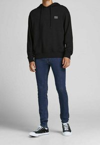 Man wearing black hoodie, blue slim-fit jeans, and black-and-white low-top sneakers, standing against a plain gray background.