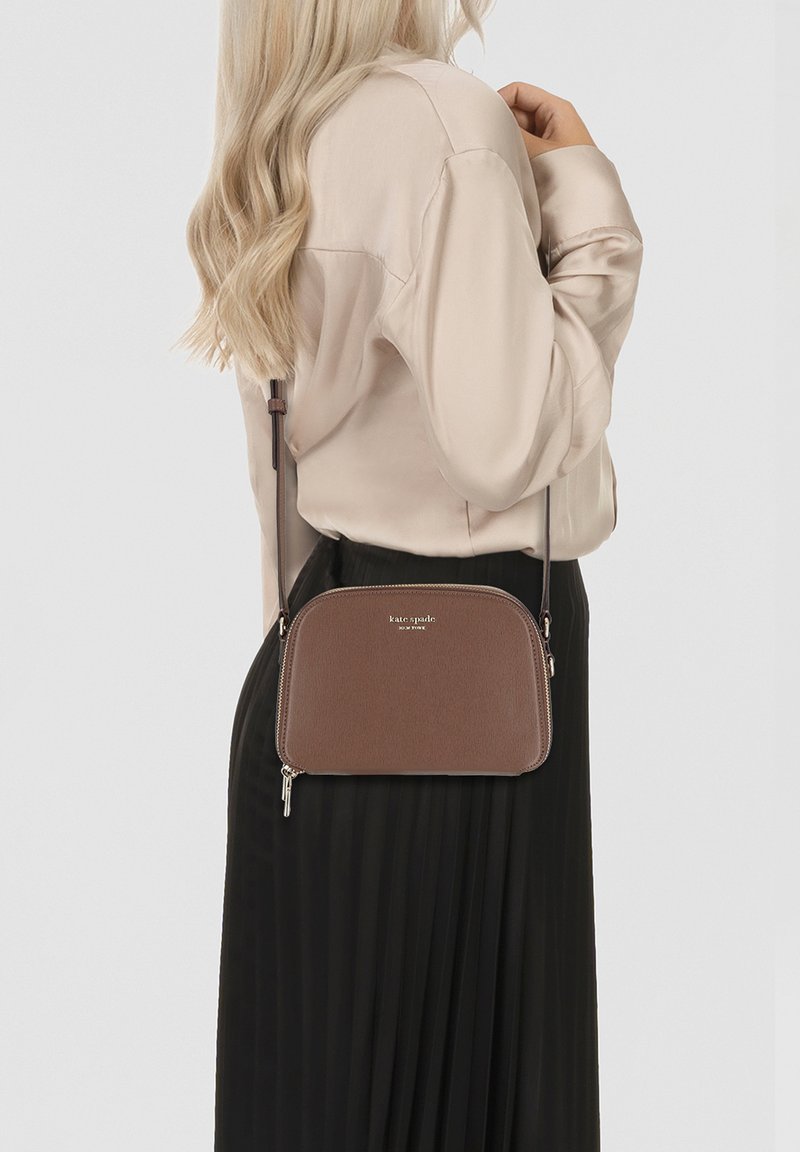 Borsa a tracolla marrone con una finitura testurizzata, forma arrotondata e chiusura con zip. Presenta accessori dorati e una tracolla, indossata su una blusa chiara.