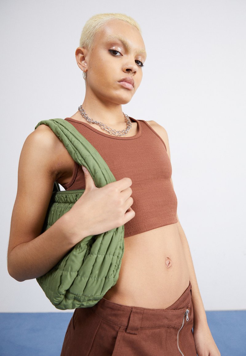Top corto marrón, bolso acolchado verde y pantalones beiges. El modelo tiene el cabello corto y rubio y lleva un collar de cadena de plata.