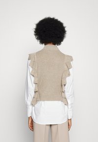 Beige neuleliivi, jossa on röyhelökoristeita, puettuna valkoisen paidan päälle. Siinä on kaulus ja istuva siluetti, jossa korostuvat teksturoidun kankaan yksityiskohdat.