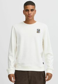 White sweatshirt with round neck and long sleeves, featuring a small embroidered "Born Lucky" patch on the left chest. Soft texture.