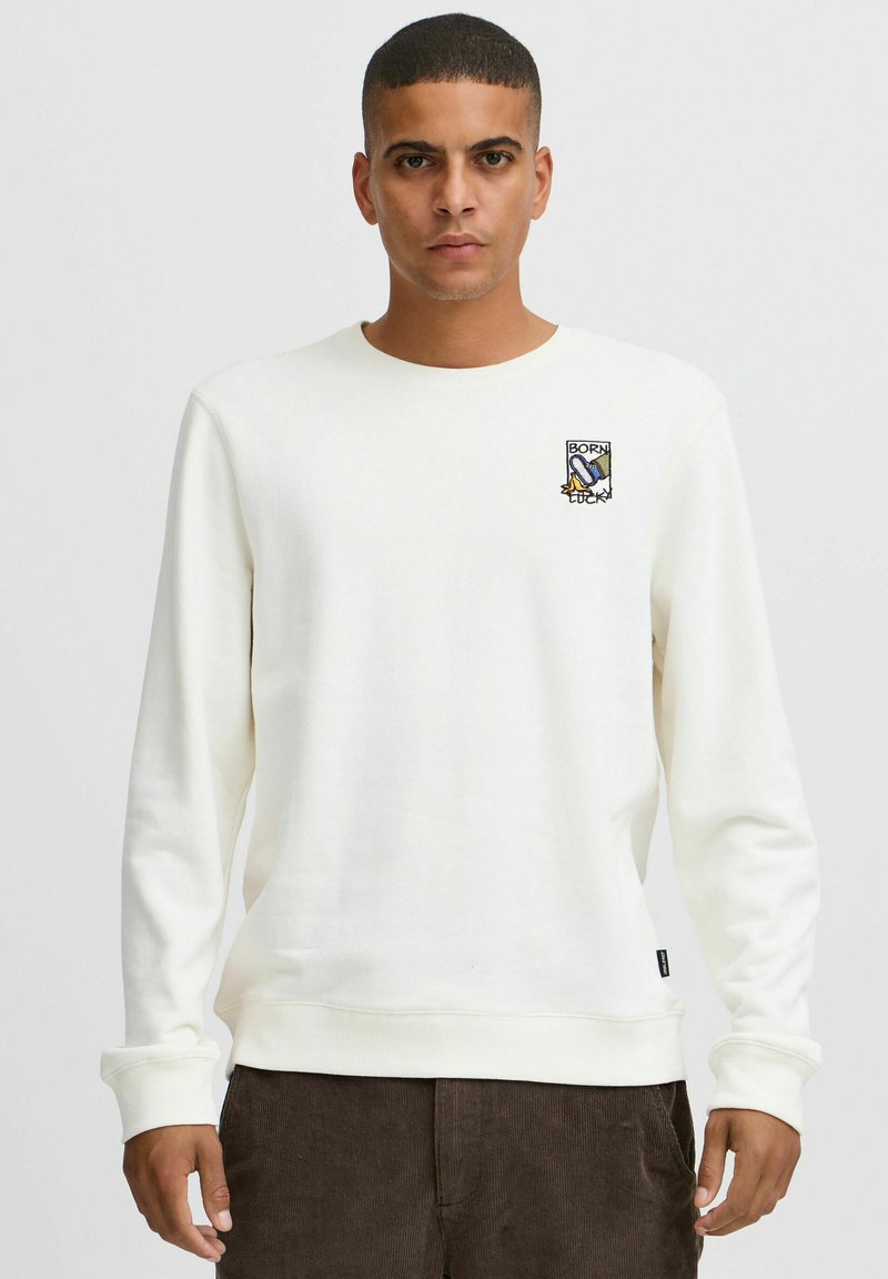 Hvit sweatshirt med rund hals og lange ermer, med et lite brodert "Born Lucky"-merke på venstre bryst. Myk tekstur.