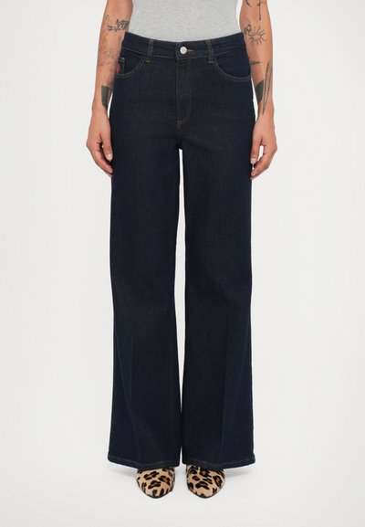 Persona vestida con pantalones de mezclilla anchos de talle alto en azul oscuro, zapatos de punta con estampado de leopardo, una blusa blanca y tatuajes visibles en los brazos.