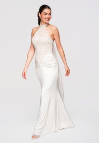 Femme portant une robe longue blanche avec un col licou en dentelle, des côtés froncés et un lien assorti en dentelle transparente au dos, souriant et regardant de côté.