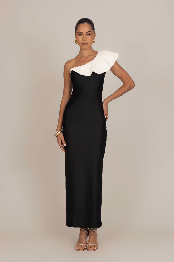 Black, fitted, one-shoulder dress with a white ruffle detail. Smooth fabric, floor-length with minimal design, paired with strappy sandals.