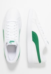 Baskets blanches avec des accents verts présentant une texture lisse, un bout arrondi, un laçage à l'avant et un logo prominent sur la languette.