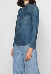 Denimskjorta i medelblått med knäppning, två bröstfickor och kontrasterande stikning. Har långa ärmar och en rundad fåll.