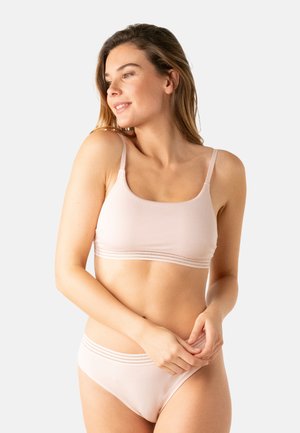 Femme à la peau claire et aux cheveux bruns portant un soutien-gorge sans couture rose pâle et un sous-vêtement assorti, debout devant un fond clair uni.