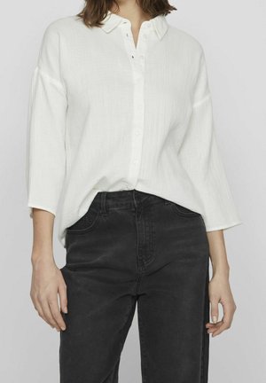 Nainen, jolla on yllään löysä valkoinen nappipaita, jossa on kolmen neljänneksen hihat, ja mustat korkeavyötäröiset farkut, tavallista taustaa vasten.